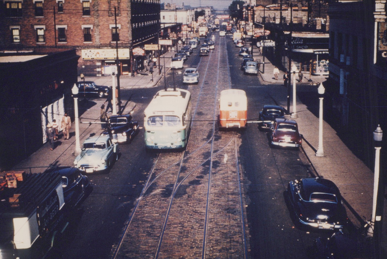 An elevated view of a street in Chicago with two trolleybuses on tracks, surrounded by classic cars and onlookers on the sidewalks.