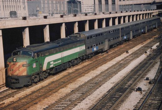 A green and white diesel locomotive numbered 9922 leads four silver Metra gallery cars on tracks beneath a concrete highway overpass, with remnants of snow visible between the rails.