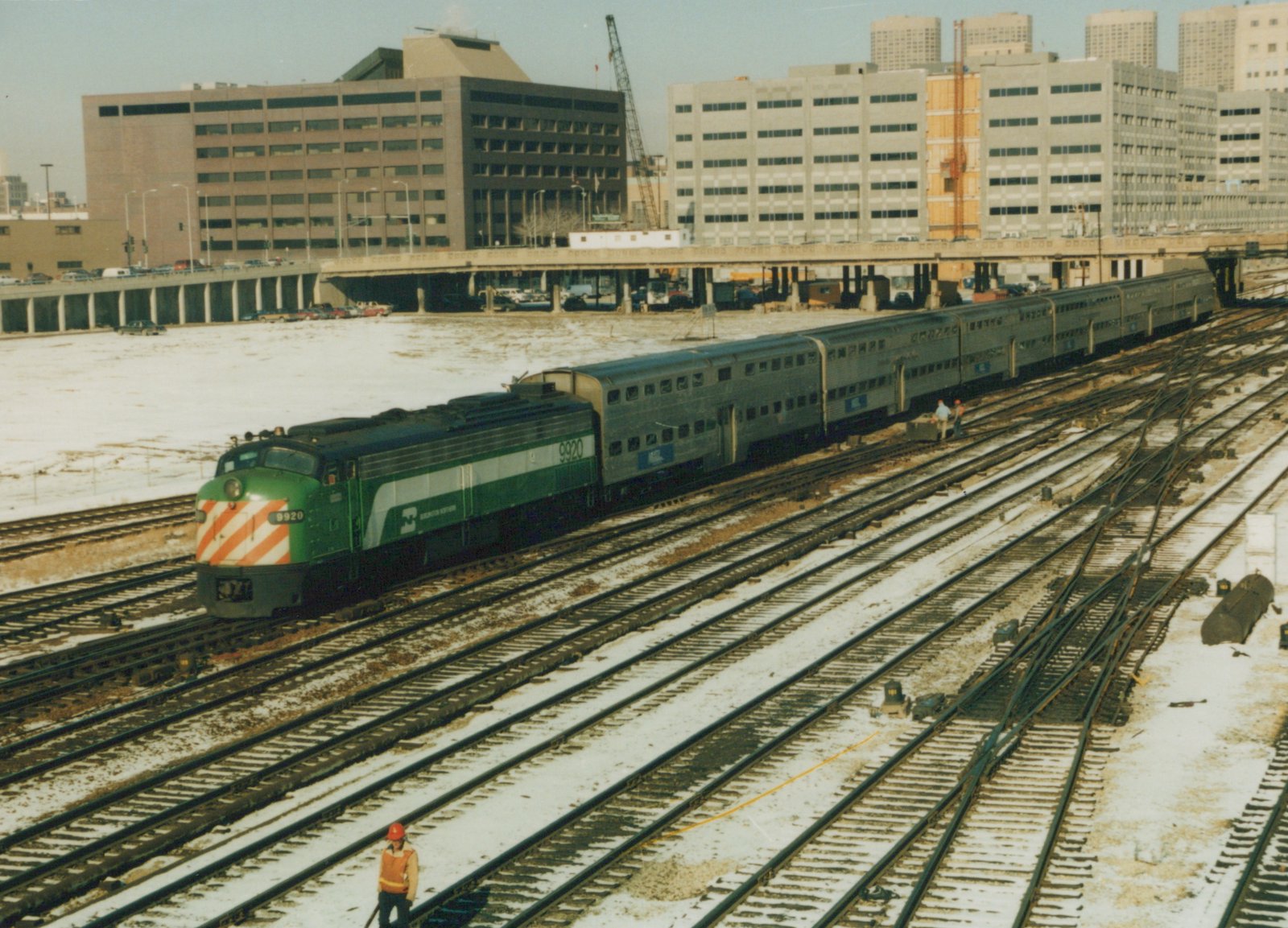 A green and white diesel locomotive with the number 9920 and 'Northwestern' lettering pulls a line of silver commuter cars through a snow-dusted rail yard surrounded by office buildings.