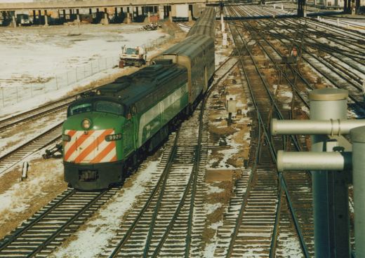 A green Burlington Northern locomotive numbered 9920 leads a train of dark passenger cars through a snowy rail yard with multiple tracks and workers visible.