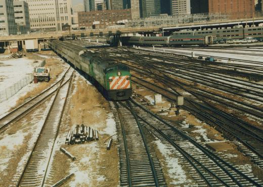 A green Metra diesel locomotive numbered 9019 travels on tracks in a winter rail yard with Amtrak cars and city buildings in the background.