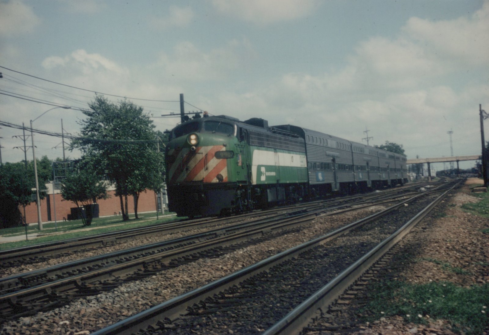 A green and white Metra diesel locomotive, number 9407, pulls a line of silver double-decker passenger cars along a set of railroad tracks next to a street with trees and a brick wall.