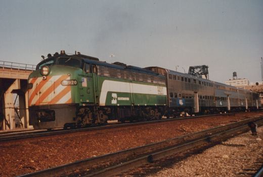 A green and white Burlington Northern diesel locomotive numbered 9920 pulls a train of silver passenger cars on railroad tracks next to a concrete bridge and a street sign reading 16TH & CANAL.