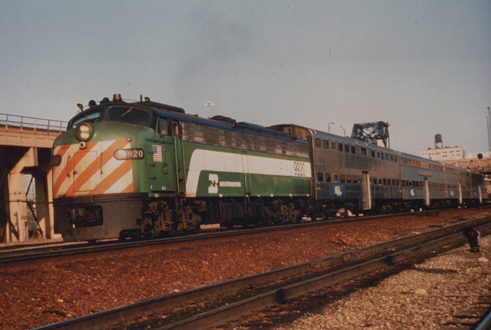 A green and white Burlington Northern diesel locomotive numbered 9920 pulls a train of silver passenger cars on railroad tracks next to a concrete bridge and a street sign reading 16TH & CANAL.