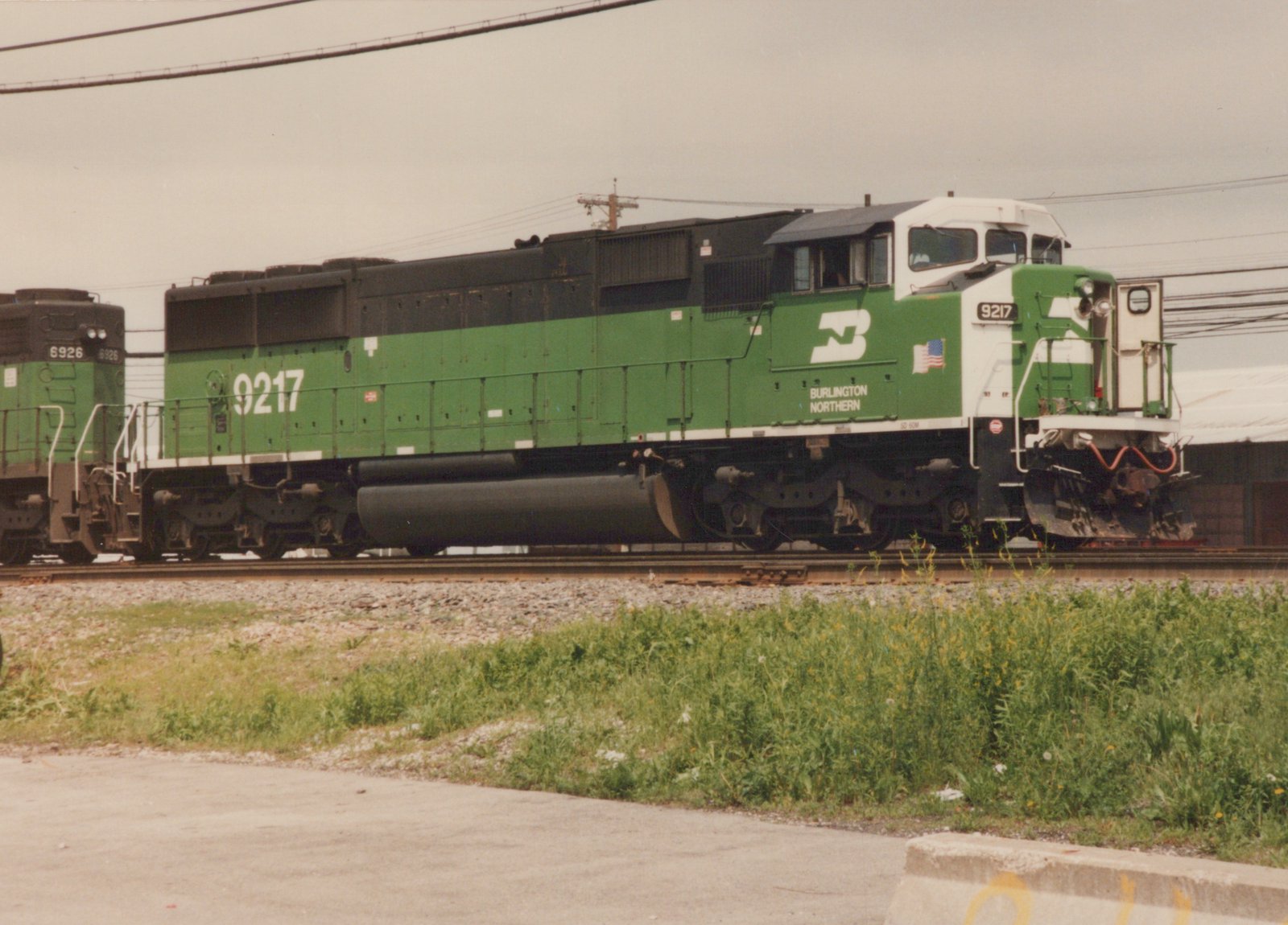 A green and white Burlington Northern locomotive numbered 9217 is shown on a clear day, with a second locomotive, number 6926, visible behind it. The train is on a private right-of-way with grass and a paved road in the foreground.