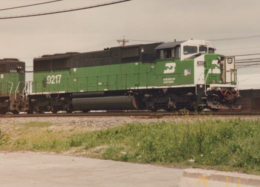 A green and white Burlington Northern locomotive numbered 9217 is shown on a clear day, with a second locomotive, number 6926, visible behind it. The train is on a private right-of-way with grass and a paved road in the foreground.