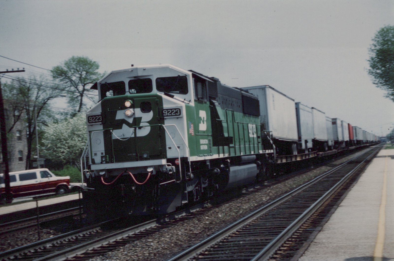 A green and white Burlington Northern locomotive numbered 9223 pulls a long line of freight cars alongside a road with a red and white station wagon parked nearby.