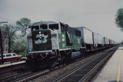A green and white Burlington Northern locomotive numbered 9223 pulls a long line of freight cars alongside a road with a red and white station wagon parked nearby.