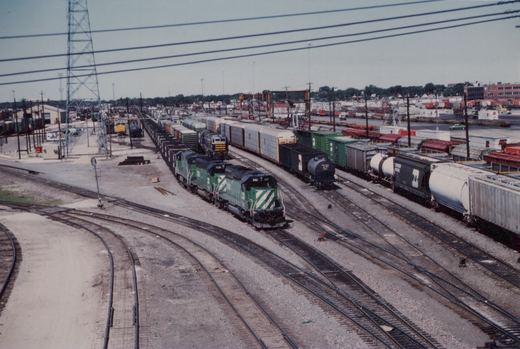An overhead view of a busy railroad yard with multiple tracks, several Burlington Northern freight trains, and industrial buildings in the background.