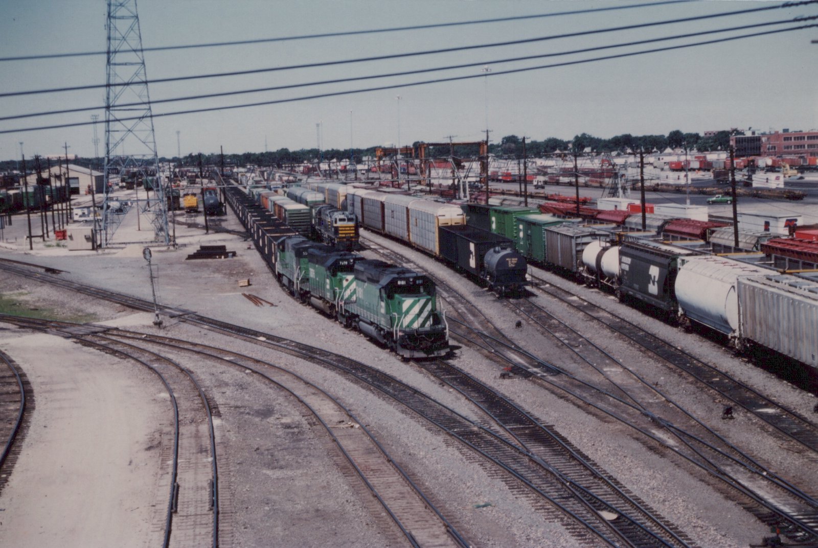 An overhead view of a busy railroad yard with multiple tracks, several Burlington Northern freight trains, and industrial buildings in the background.