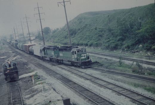 A green and white Norfolk and Western locomotive numbered 8054 pulls a long freight train on multiple tracks in a grassy, hilly area under a cloudy sky.
