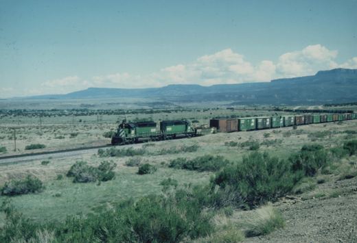 Two green Burlington Northern locomotives pulling a long freight train through a dry, brush-covered landscape with distant mountains under a blue sky with white clouds.