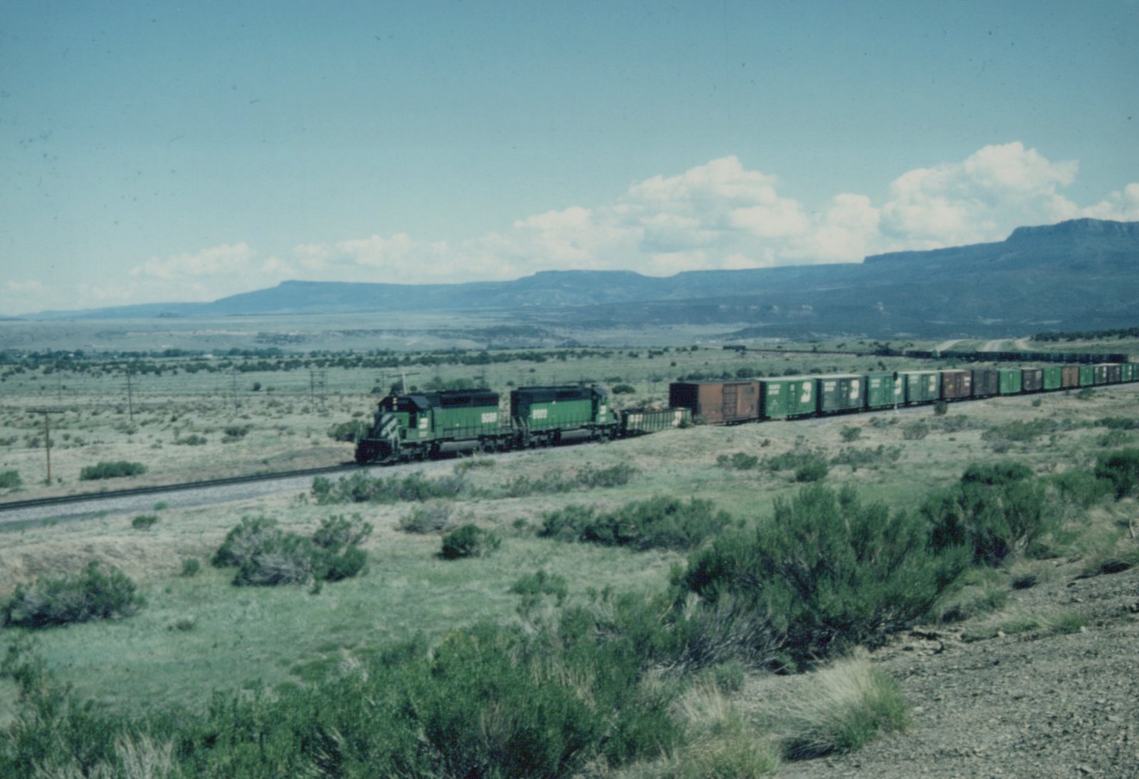 Two green Burlington Northern locomotives pulling a long freight train through a dry, brush-covered landscape with distant mountains under a blue sky with white clouds.