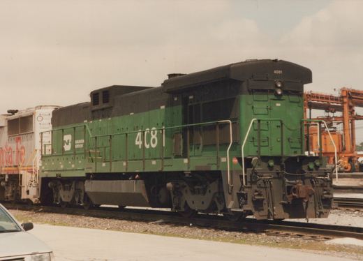 A green and black Burlington Northern GP38-2 diesel locomotive numbered 4081 sits on tracks in a rail yard, with another locomotive and a car visible in the background.