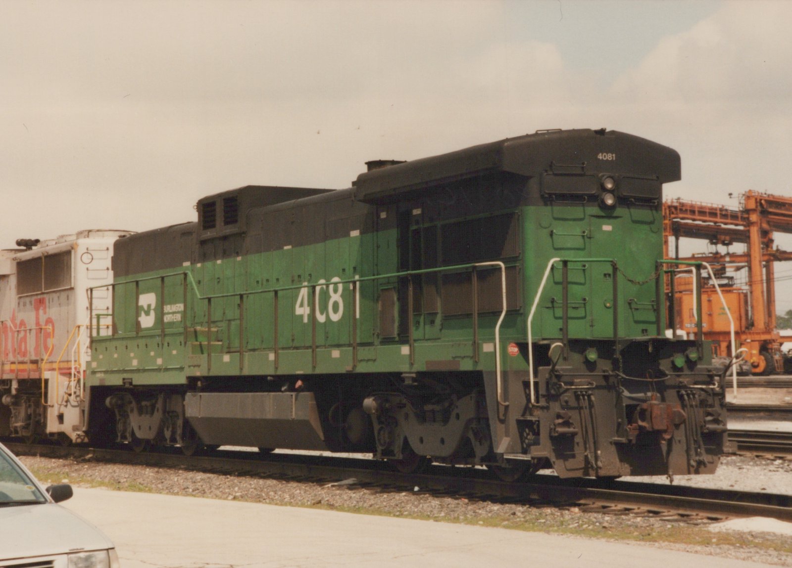 A green and black Burlington Northern GP38-2 diesel locomotive numbered 4081 sits on tracks in a rail yard, with another locomotive and a car visible in the background.