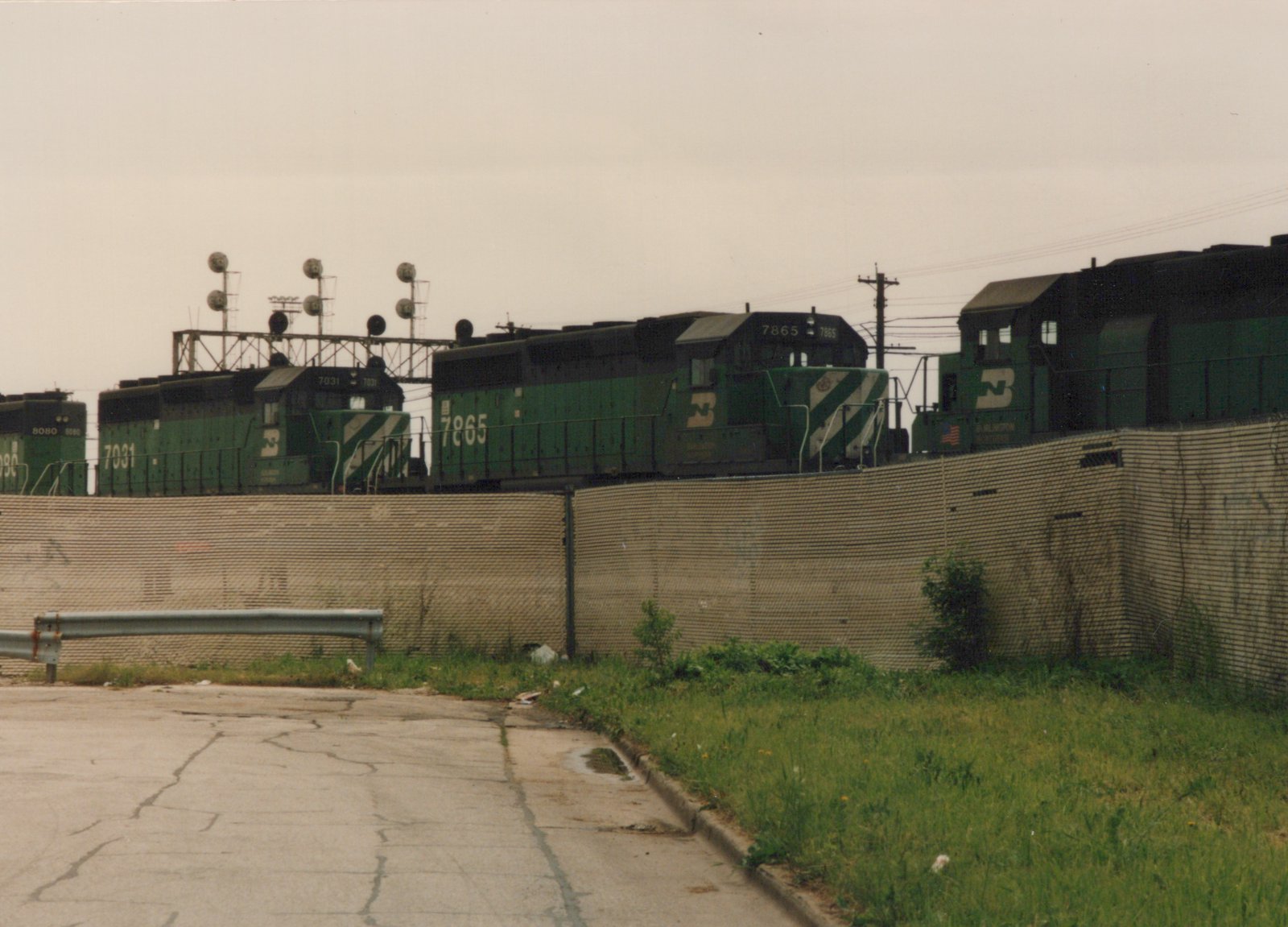 Three green Burlington Northern diesel locomotives, numbers 7865, 7031, and 8080, are on tracks behind a chain-link fence and a concrete wall, with cracked pavement in the foreground.