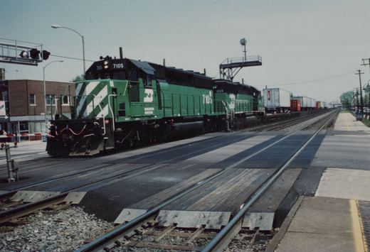 Two green Burlington Northern SD40-2 diesel locomotives, numbers 7105 and 3149, pull a freight train across a grade crossing on a cloudy day.