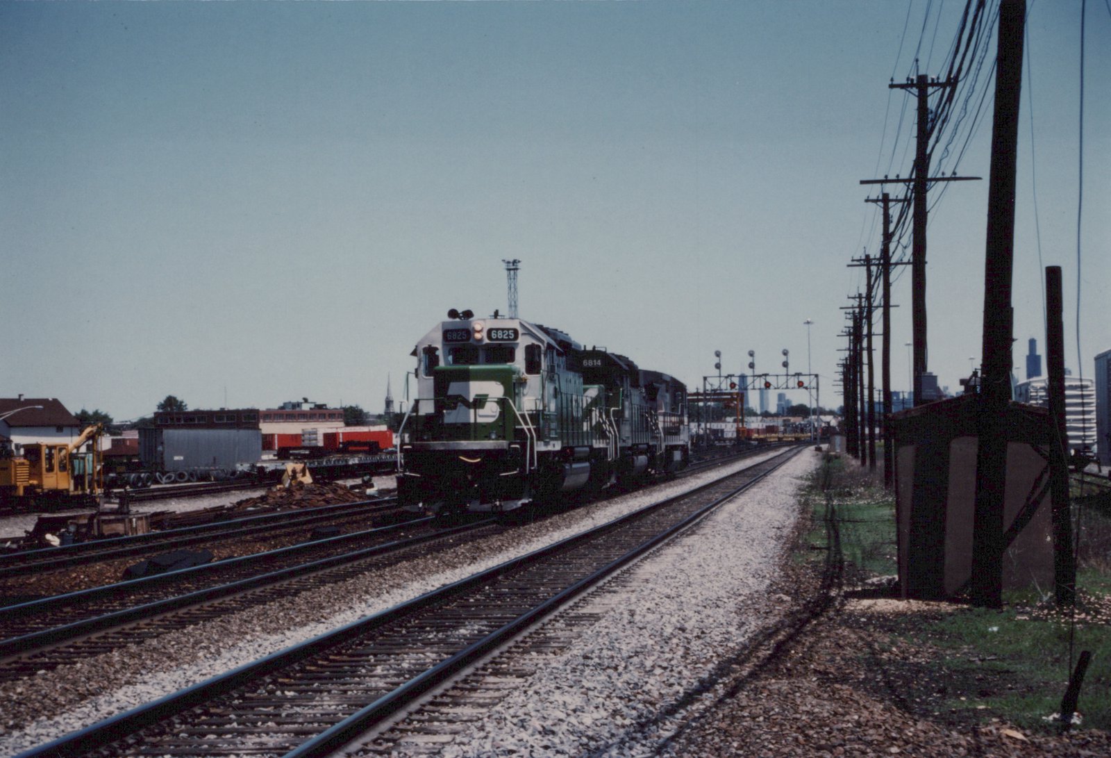A diesel locomotive painted in Burlington Northern's green and white livery leads a freight train on a sunny day, with a city skyline in the background and power lines running alongside the tracks.