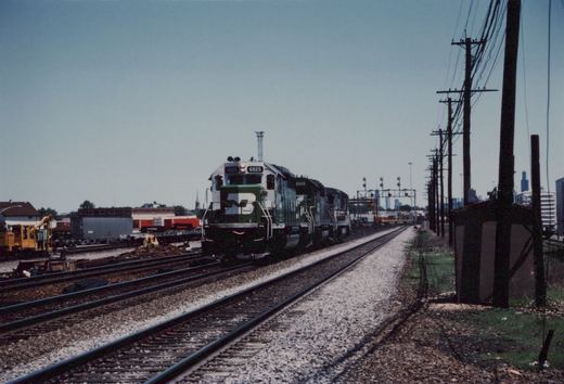 A diesel locomotive painted in Burlington Northern's green and white livery leads a freight train on a sunny day, with a city skyline in the background and power lines running alongside the tracks.