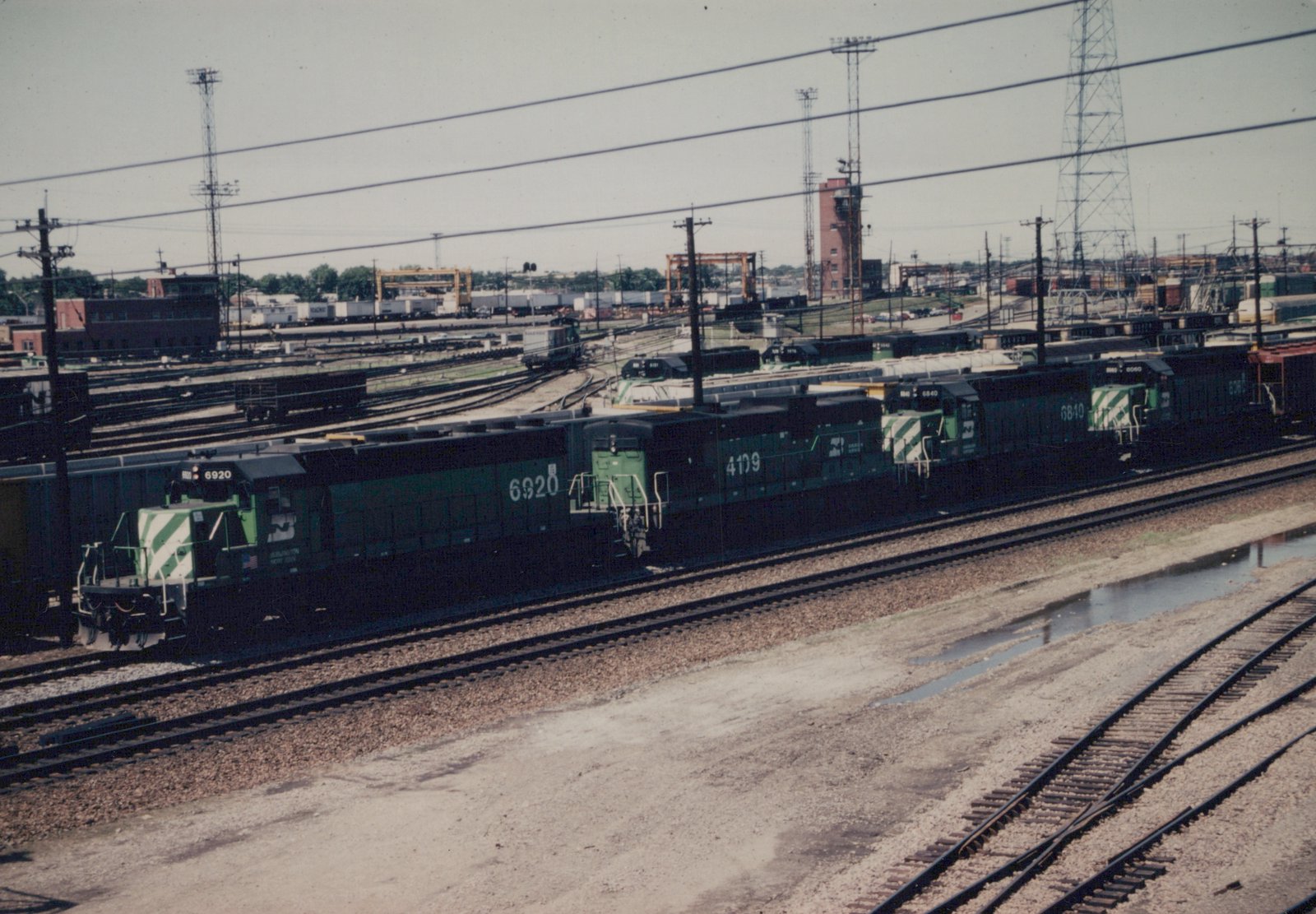 Four green Burlington Northern diesel locomotives are lined up on parallel tracks in a large railroad yard with industrial buildings and cranes visible in the background on a clear day.