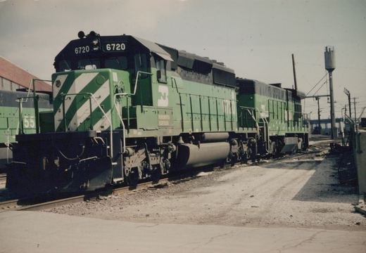 Three green Burlington Northern diesel locomotives, including 6720 in the lead, are parked on tracks in a gravel yard with a water tower in the background.