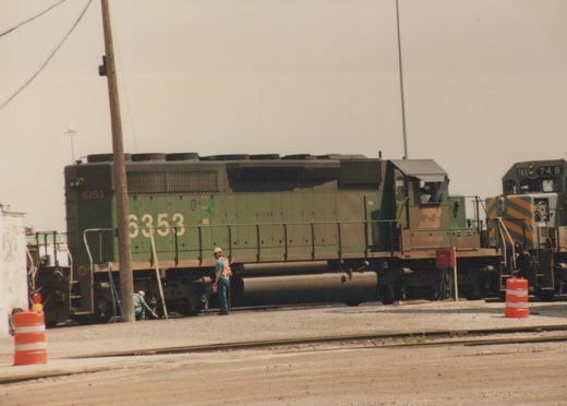 A green Burlington Northern diesel locomotive numbered 6353 is parked in a rail yard with two workers nearby, and another locomotive numbered 748 is visible behind it.