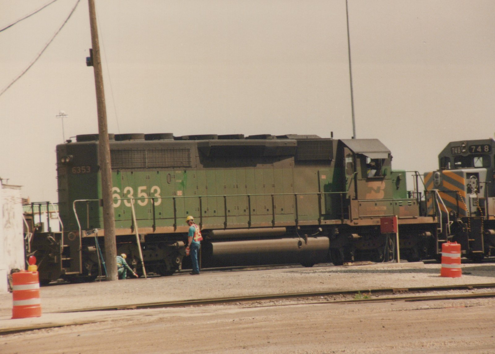 A green Burlington Northern diesel locomotive numbered 6353 is parked in a rail yard with two workers nearby, and another locomotive numbered 748 is visible behind it.