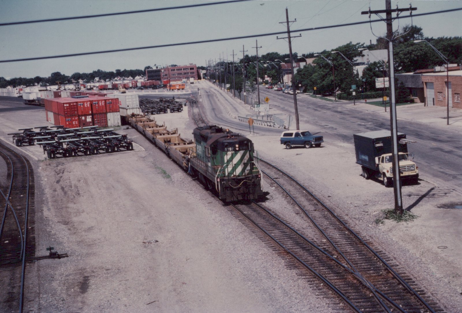 A green and white Milwaukee Road diesel locomotive, number 6197, sits on tracks in a rail yard, with trucks and shipping containers nearby.