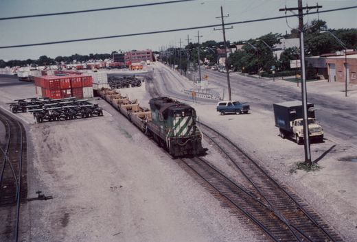 A green and white Milwaukee Road diesel locomotive, number 6197, sits on tracks in a rail yard, with trucks and shipping containers nearby.