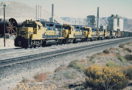 A long Santa Fe freight train with several blue and yellow diesel locomotives leads a line of freight cars through a dry, shrub-covered landscape with industrial buildings and hills in the background.