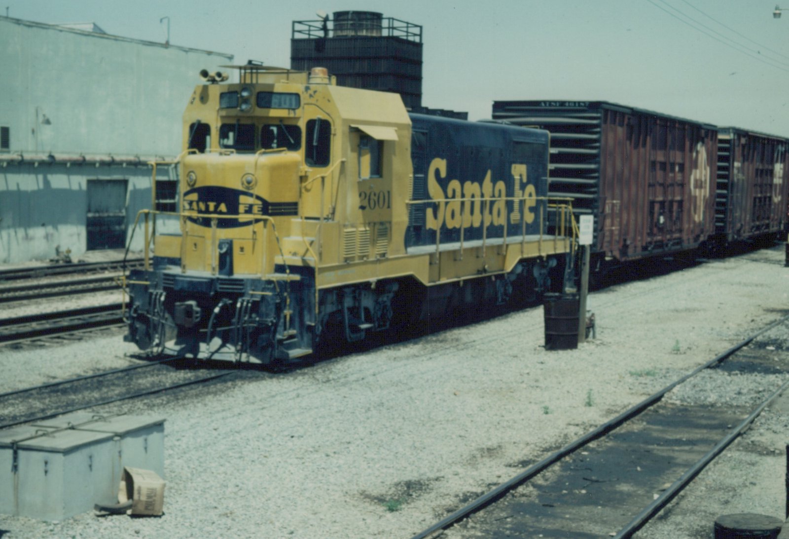 A yellow and blue Santa Fe locomotive numbered 2601 is coupled to several freight cars in a gravel rail yard on a clear day.