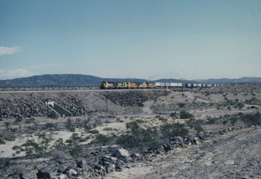 A long freight train with yellow and black locomotives moves across a desert landscape with rocky terrain and sparse shrubs, mountains visible in the distance.