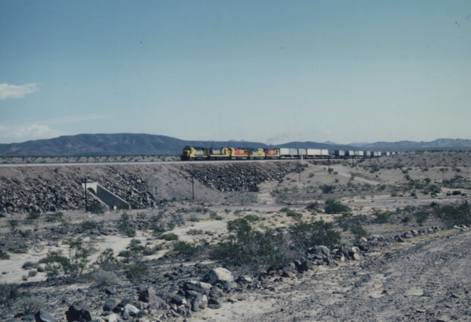 A long freight train with yellow and black locomotives moves across a desert landscape with rocky terrain and sparse shrubs, mountains visible in the distance.