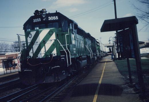 A green and white Burlington Northern diesel locomotive, number 3055, is at a station platform with bare trees and a small shelter beside it.