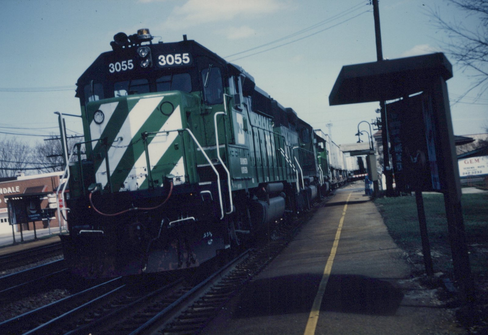 A green and white Burlington Northern diesel locomotive, number 3055, is at a station platform with bare trees and a small shelter beside it.