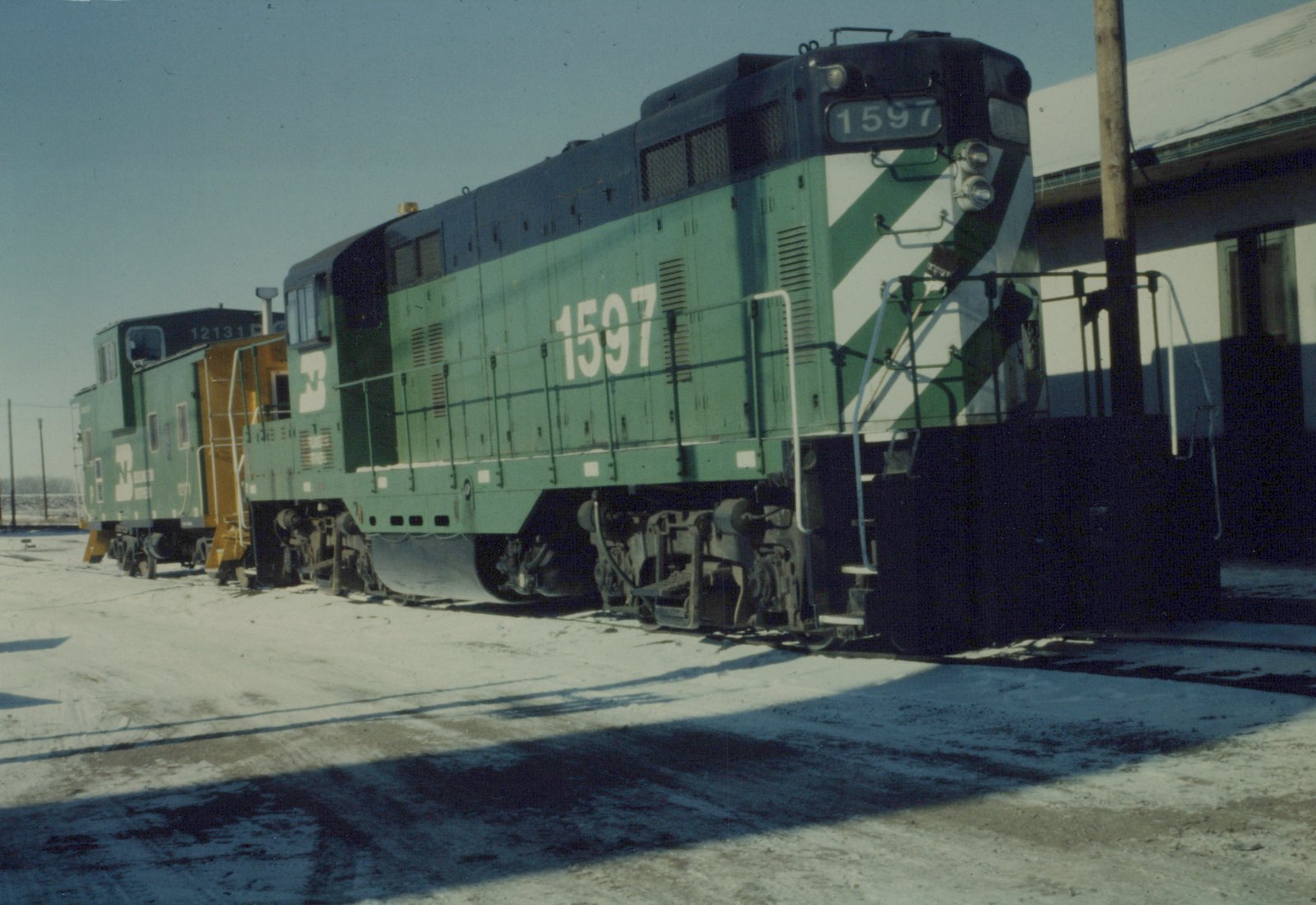 A green and white Burlington Northern diesel locomotive number 1597 is parked on a snowy railroad yard with a caboose behind it.