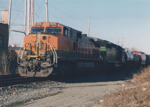 A bright orange and black BNSF freight train with two locomotives and tank cars travels on tracks beside a gravel shoulder and dry grass on a sunny, clear day.