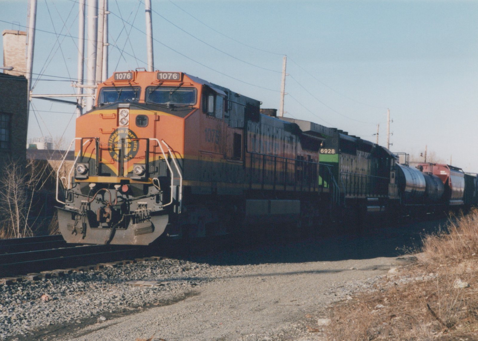 A bright orange and black BNSF freight train with two locomotives and tank cars travels on tracks beside a gravel shoulder and dry grass on a sunny, clear day.