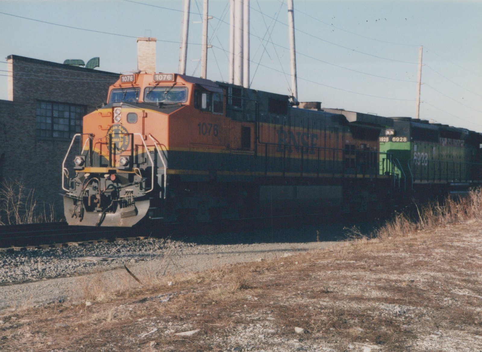 A three-unit BNSF diesel locomotive consist, led by locomotive number 1076, travels along tracks bordered by dry grass and a brick building under a clear sky.