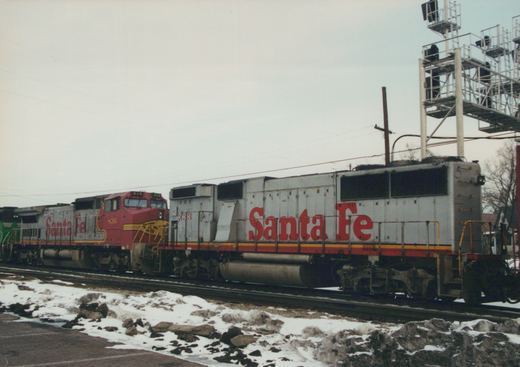 A side view of two Santa Fe freight locomotives, one red and one silver, with snow on the ground.