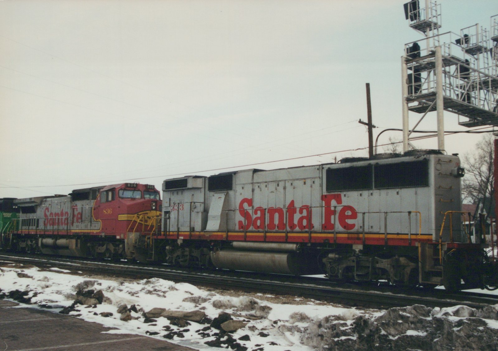 A side view of two Santa Fe freight locomotives, one red and one silver, with snow on the ground.