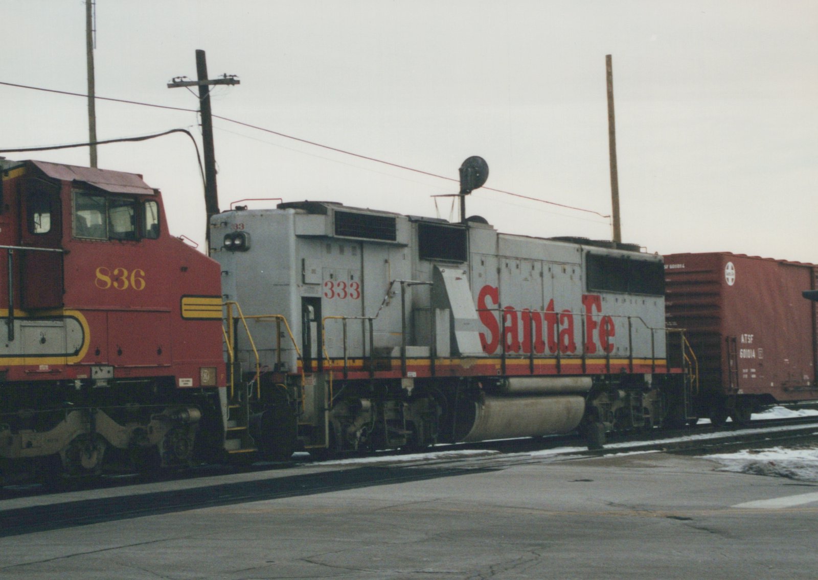 A red and a gray Santa Fe GP9 diesel locomotive side by side in a snowy train yard, with freight cars behind and snow on the ground.