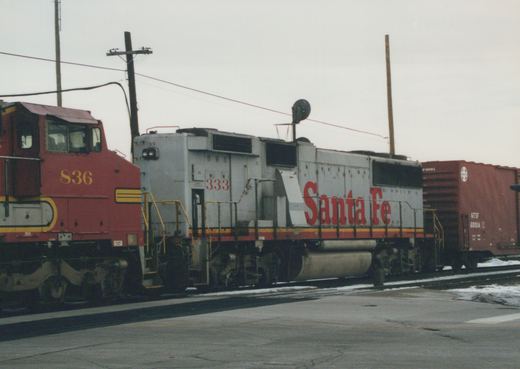 A red and a gray Santa Fe GP9 diesel locomotive side by side in a snowy train yard, with freight cars behind and snow on the ground.