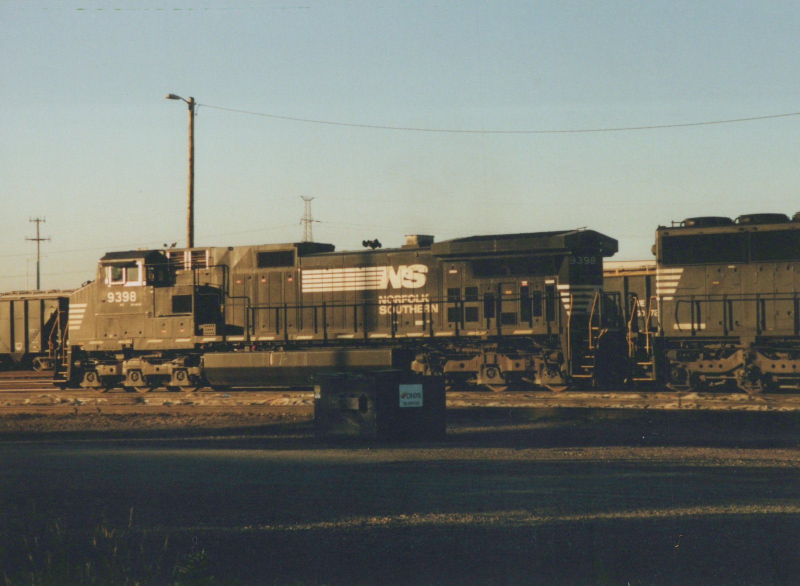 Side view of a dark gray Norfolk Southern diesel locomotive, number 9398, with white lettering, parked on tracks in a yard on a clear day.