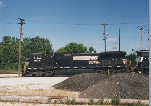 Side view of two black Norfolk Southern diesel locomotives, numbers 9219 and 2729, parked in a rail yard with trees and utility poles in the background.