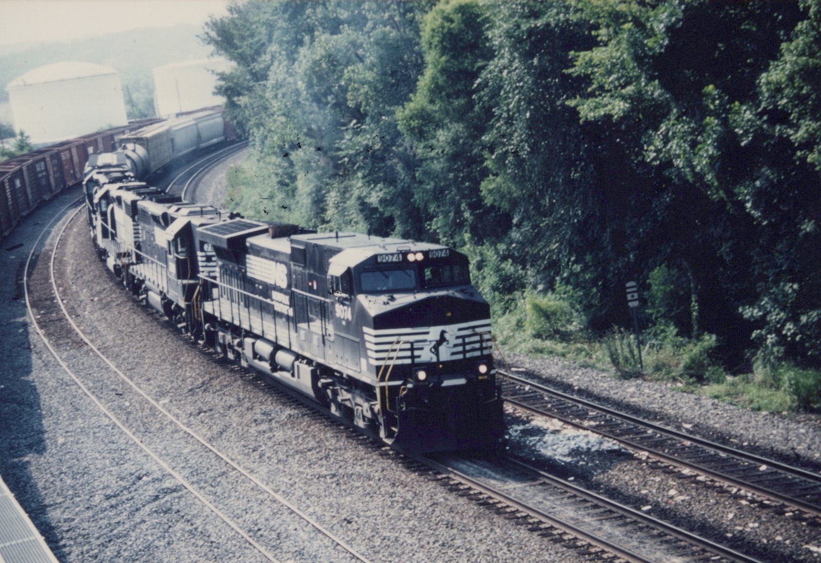 A black and white Norfolk Southern freight train, led by locomotive 9074, curves along tracks bordered by green trees and industrial buildings.