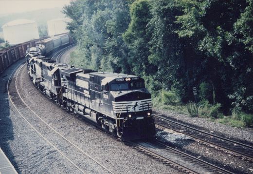 A black and white Norfolk Southern freight train, led by locomotive 9074, curves along tracks bordered by green trees and industrial buildings.