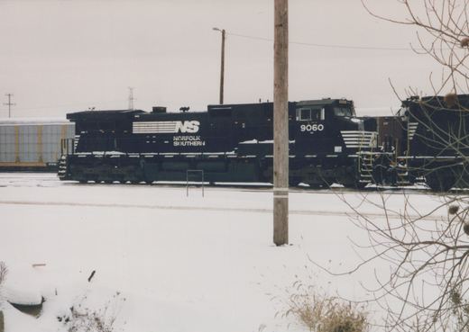 A black Norfolk Southern locomotive with the number 9060 is parked on railroad tracks in a snow-covered yard, with a freight car visible in the background.