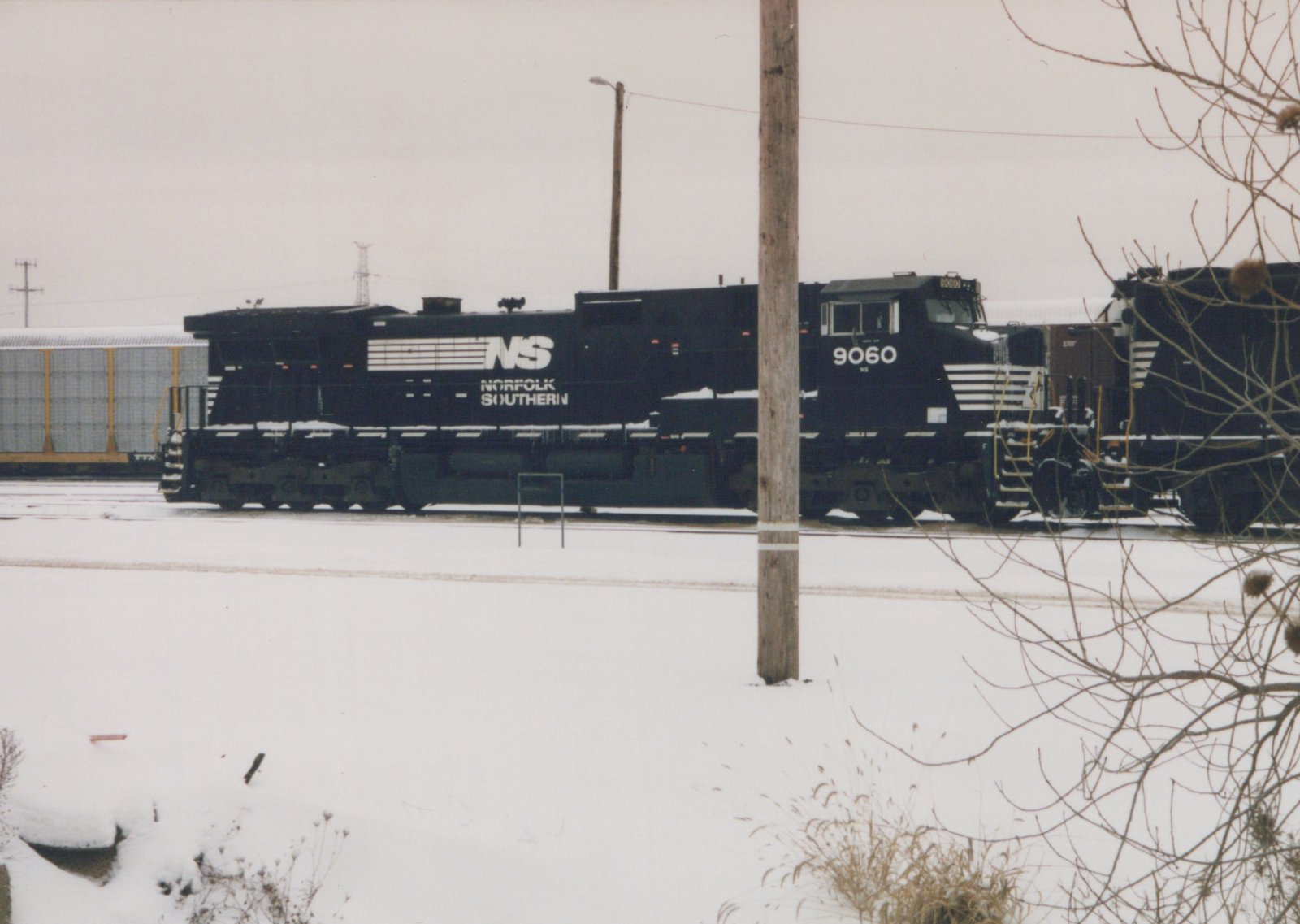 A black Norfolk Southern locomotive with the number 9060 is parked on railroad tracks in a snow-covered yard, with a freight car visible in the background.