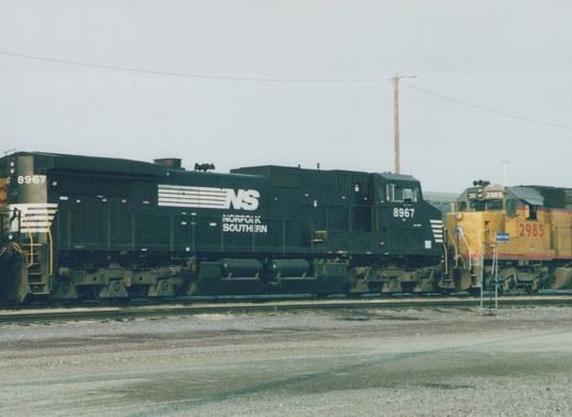 Side view of a black Norfolk Southern locomotive numbered 8967 in a railyard, with a yellow locomotive numbered 2985 on an adjacent track under an overcast sky.